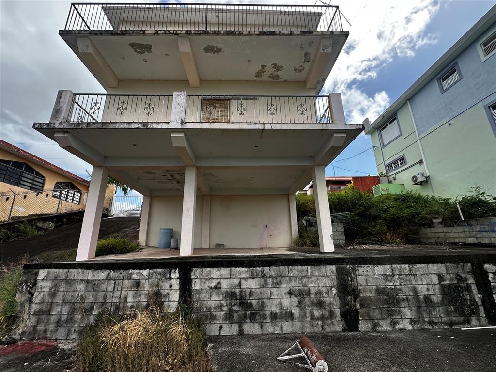 A two-story house with an open ground level, concrete balconies, and a stone retaining wall — D URB. EL EDEN D-7, COAMO, PR