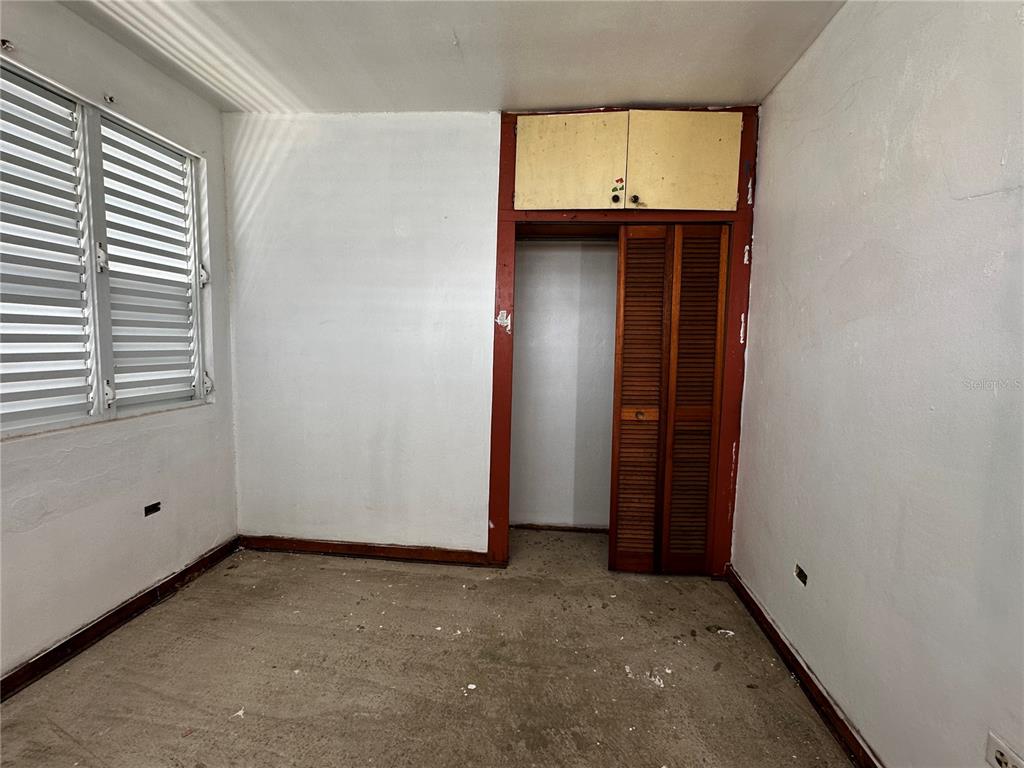 An empty room features white walls, a louvered window, and a closet with wooden doors — D URB. EL EDEN D-7, COAMO, PR