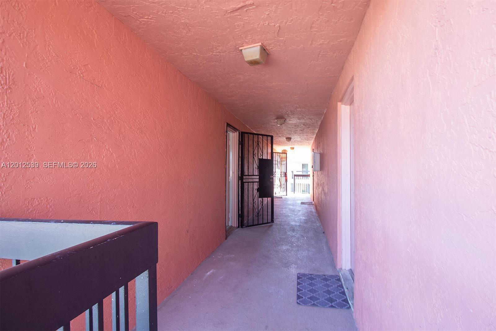An outdoor apartment hallway features pink stucco walls, a concrete floor, and metal security doors — 2020 NW 119th St # 1121, Miami FL 33167, MIAMI, FL