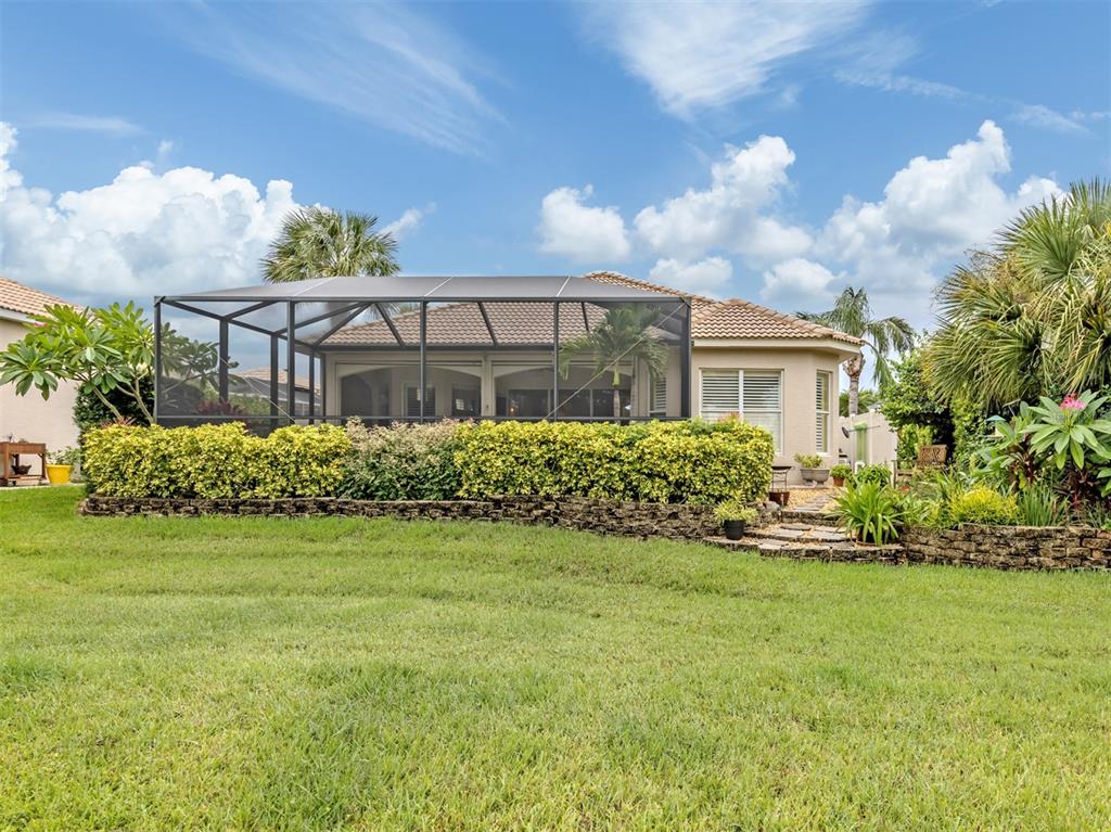 A backyard view of a house with a screened-in lanai and a stone retaining wall — 151 COCO PALM DRIVE, VENICE, FL