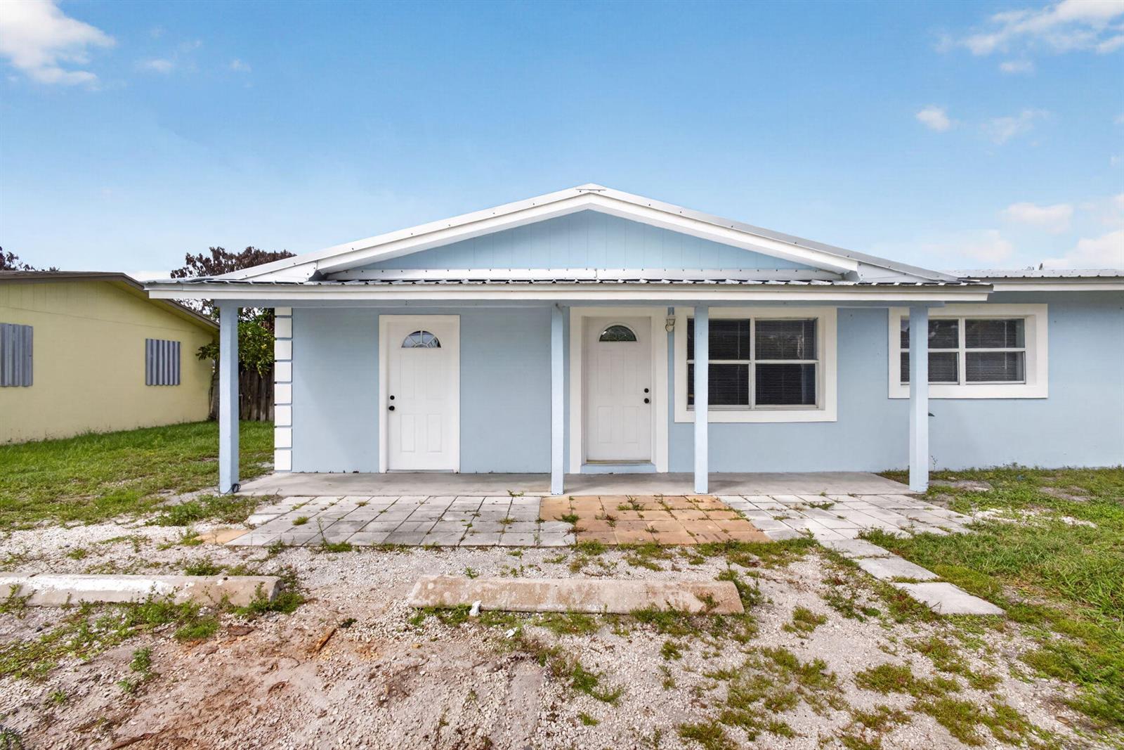 A light blue single-story house features a covered porch, two white doors, and a gravel yard — 15478 SW 150th Street, FL 34956, FL
