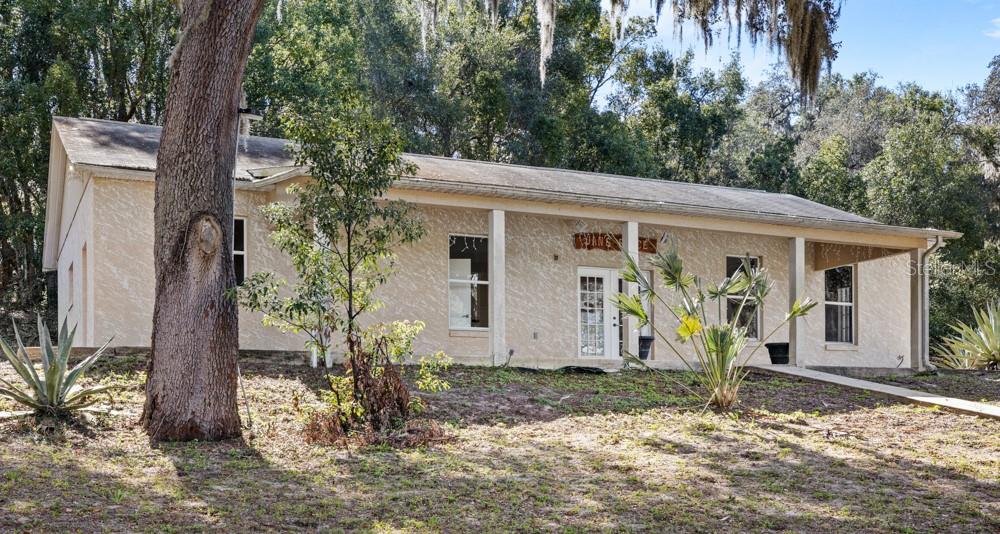 A single-story, light-colored stucco house with a covered porch sits on a wooded lot — 19621 LAKE LINCOLN LANE, EUSTIS, FL