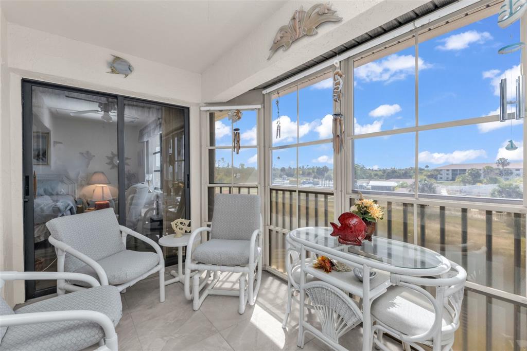 A sunroom features white wicker furniture, tile flooring, and large windows overlooking a grassy landscape — 175 KINGS HIGHWAY UNIT 1032, PUNTA GORDA, FL