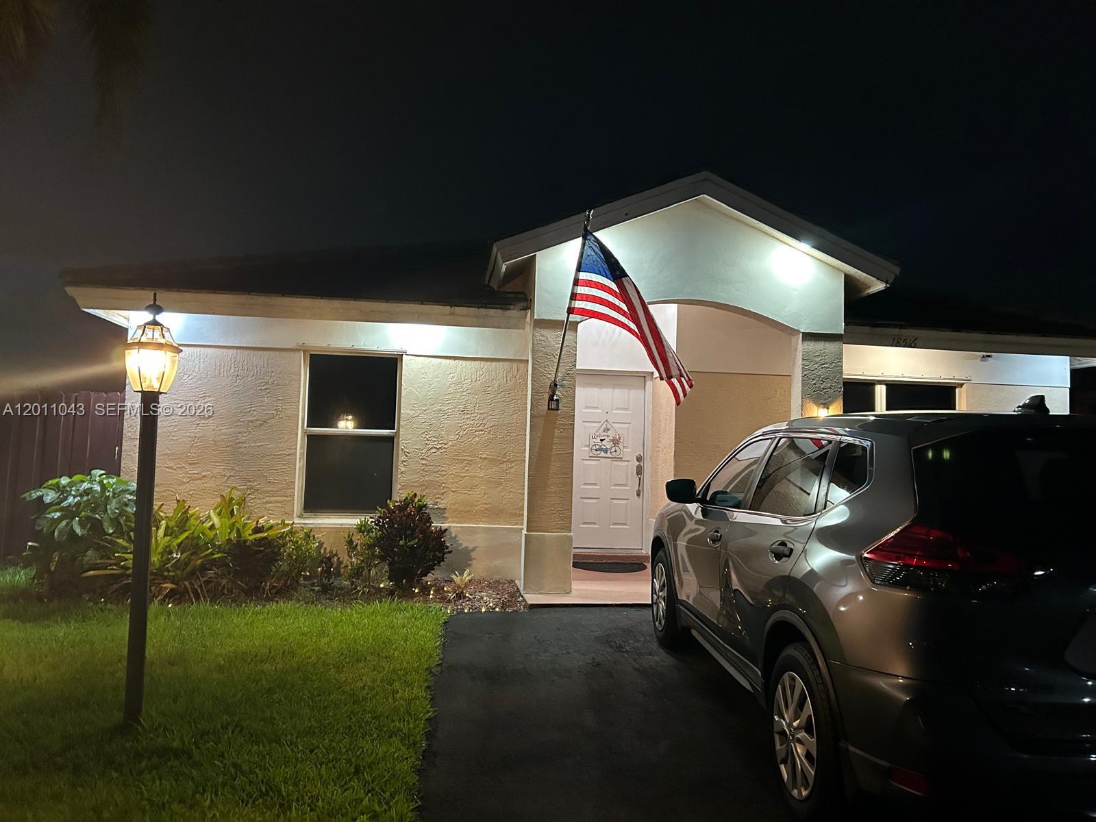 A single-story house exterior at night with a parked car and an American flag — 18516 NW 56th Ave, Miami Gardens FL 33055, MIAMI GARDENS, FL