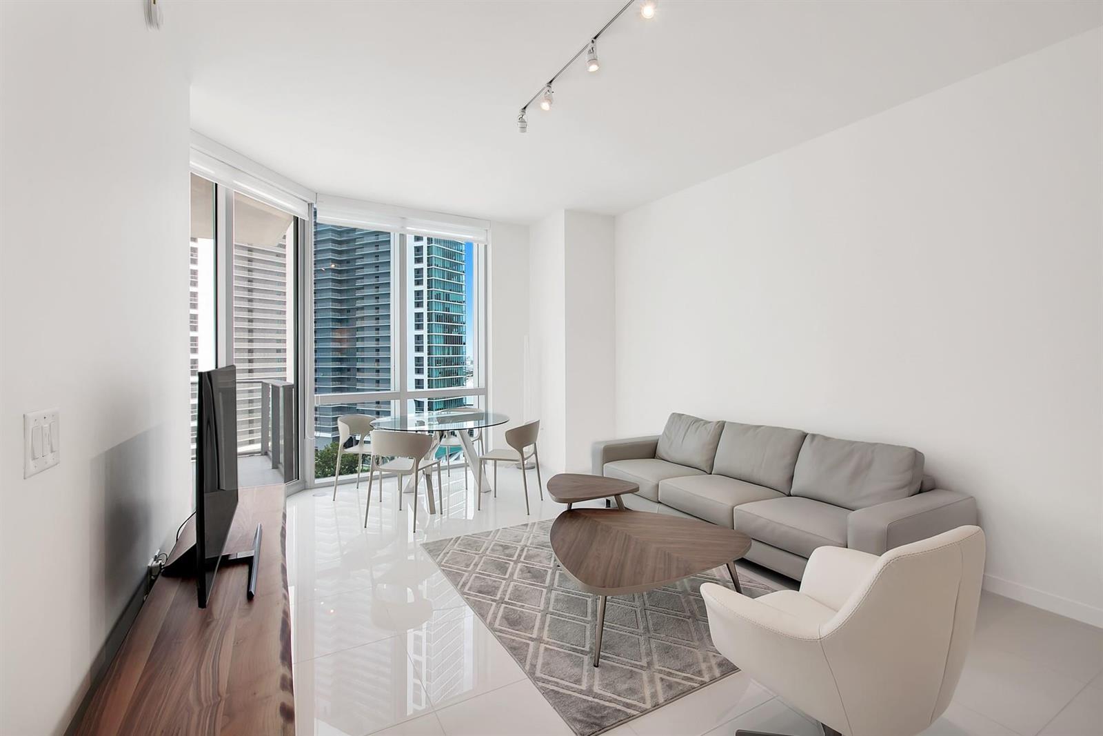 A modern living room with a gray sofa, white armchair, glass dining table, and city views — 851 NE 1st Avenue # 2005, Miami FL 33132, MIAMI, FL