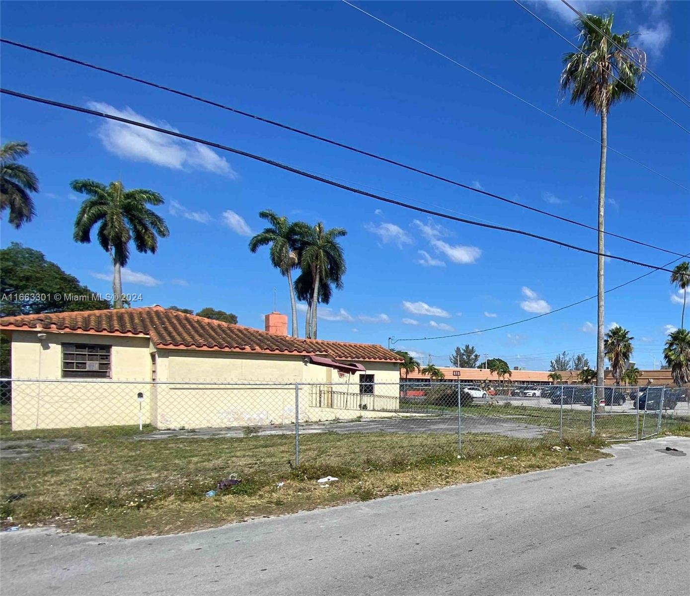 A single-story building with a tiled roof sits behind a chain-link fence near palm trees — 13400 Alexandria Dr, Opa-Locka FL 33054, OPA-LOCKA, FL