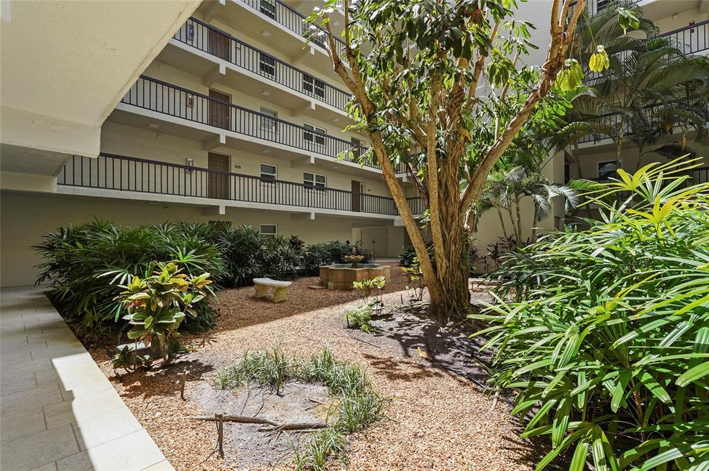 An outdoor courtyard features a central fountain, gravel ground, lush landscaping, and multi-story apartment balconies — 3240 GULF OF MEXICO DRIVE UNIT B104, LONGBOAT KEY, FL