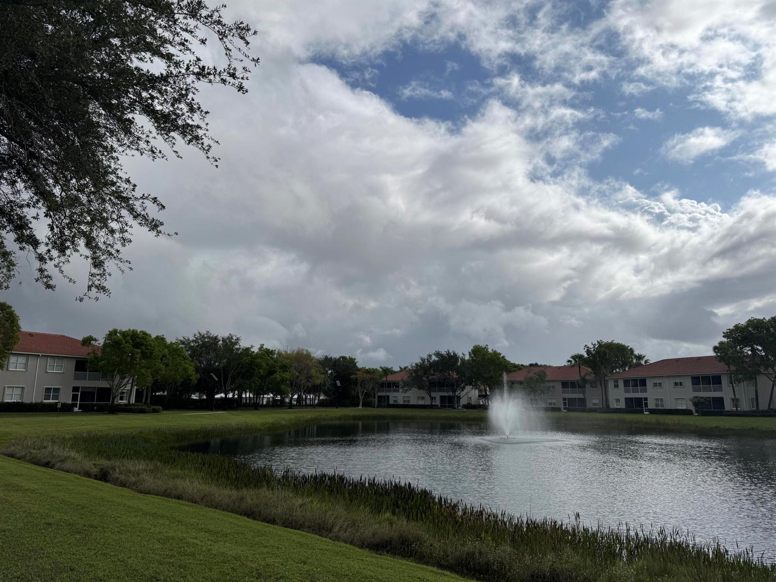 A pond with a fountain sits in front of apartment buildings under a cloudy sky — 2140 Wingate Bend, Wellington FL 33414, WELLINGTON, FL