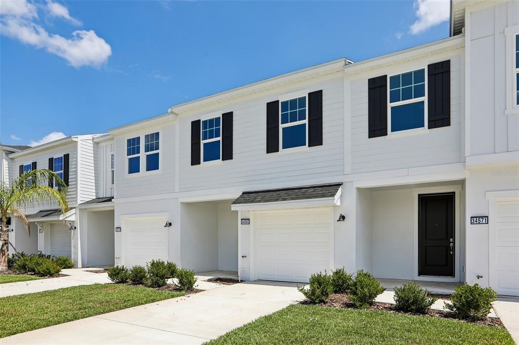 A row of white two-story townhouses with black shutters and attached garages under a blue sky — 34563 TURPINS AVENUE, ZEPHYRHILLS, FL