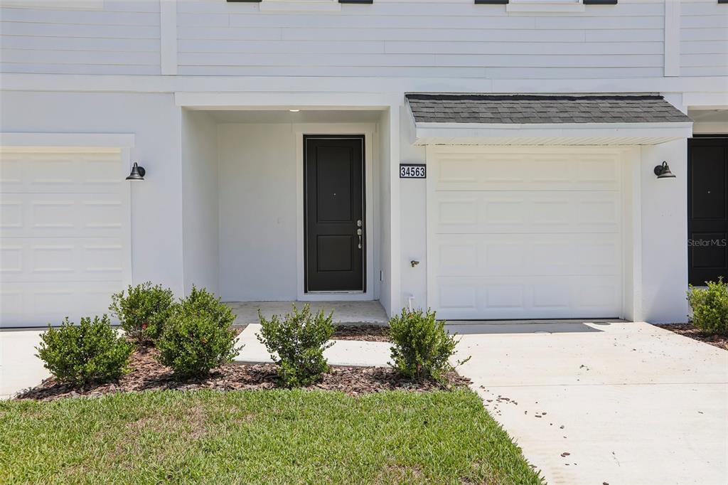 A white townhouse exterior features a black front door, a garage, and small green shrubs — 34563 TURPINS AVENUE, ZEPHYRHILLS, FL