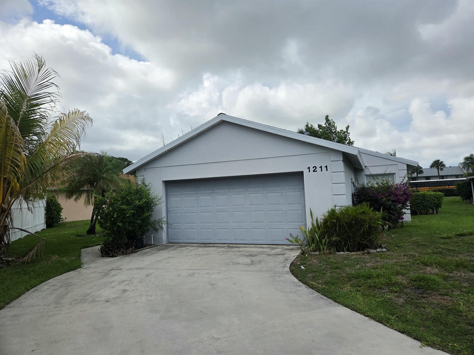 A single-story house with a gray garage door and a concrete driveway under a cloudy sky — 1211 Pine Sage Circle, West Palm Beach FL 33409, WEST PALM BEACH, FL