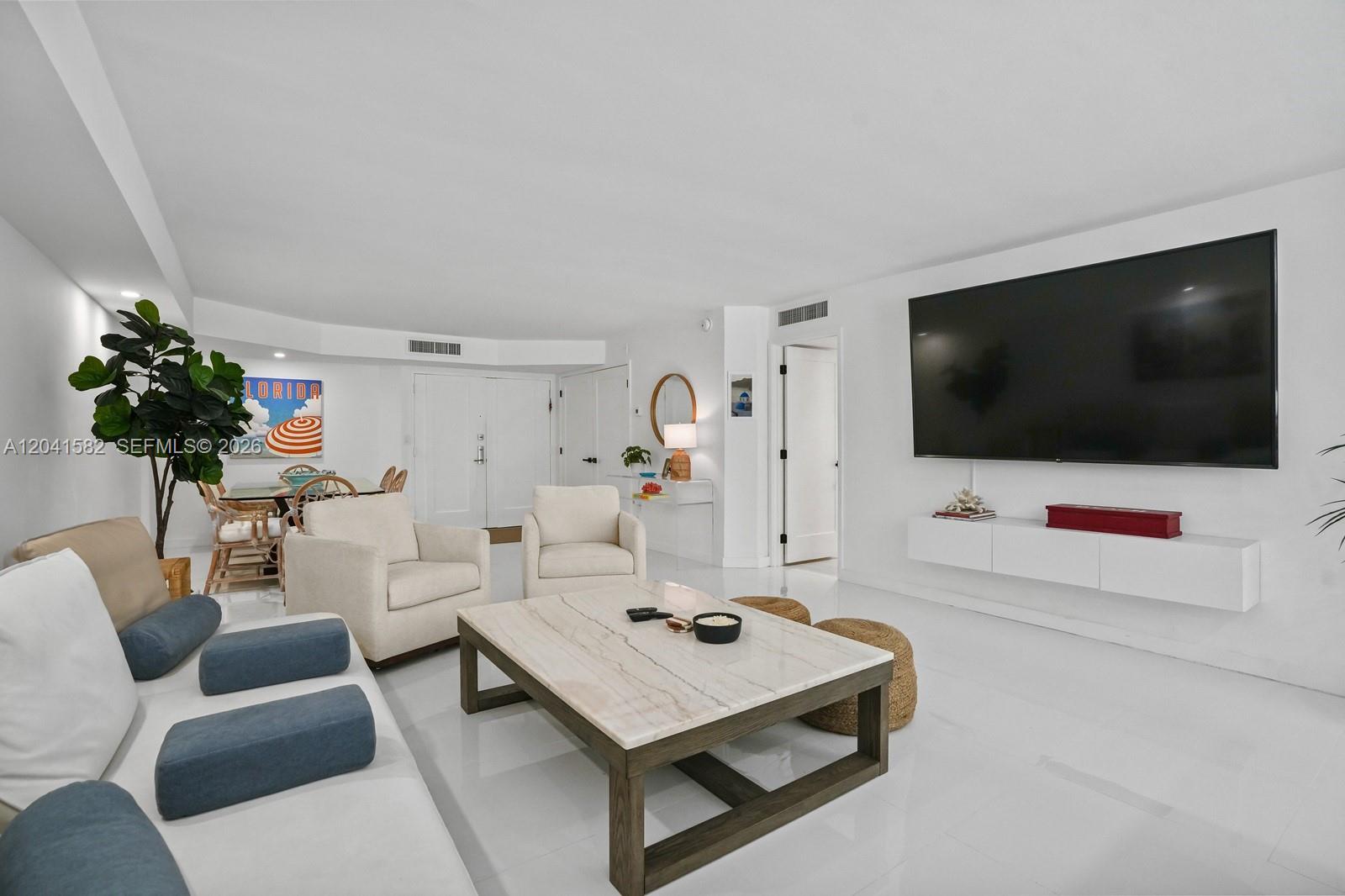 A bright living room features white flooring, a large wall-mounted television, and neutral furniture — 6423 Collins Ave # 203, Miami Beach FL 33141, MIAMI BEACH, FL