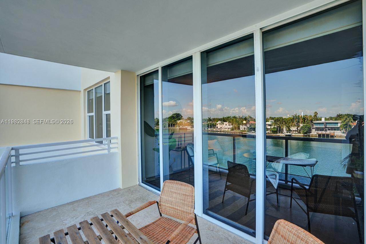 A balcony with wicker chairs overlooks a waterfront view through large sliding glass doors — 5600 Collins Ave # 5R, Miami Beach FL 33140, MIAMI BEACH, FL