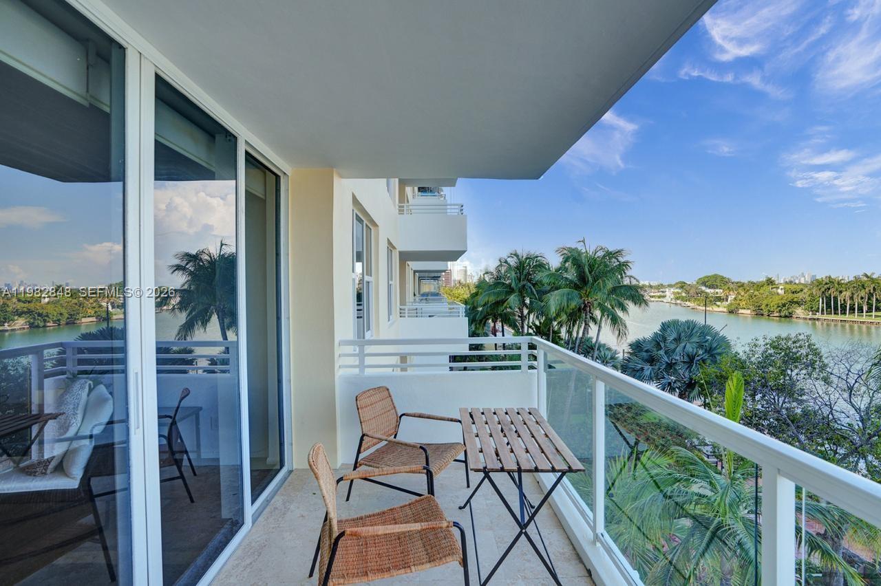 A balcony with wicker chairs and a wooden table overlooks a waterway and palm trees — 5600 Collins Ave # 5R, Miami Beach FL 33140, MIAMI BEACH, FL