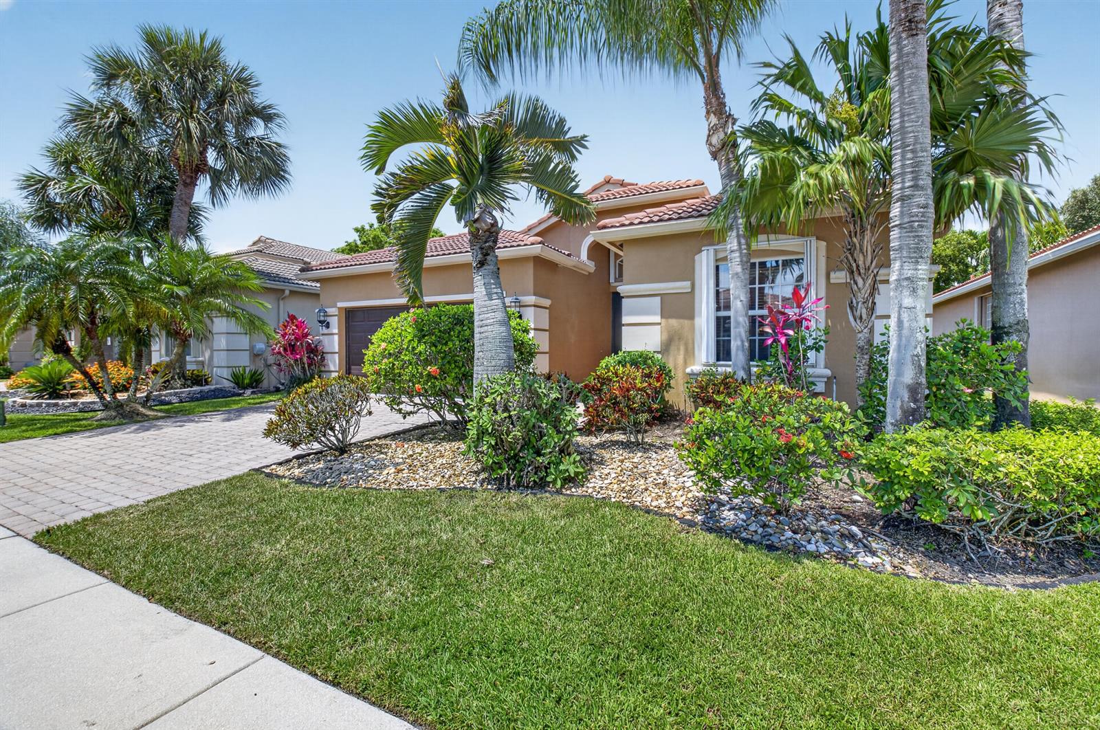 A single-story house with a paved driveway, palm trees, and landscaped garden under a blue sky — 6761 Pisano Drive, Lake Worth FL 33467, LAKE WORTH, FL