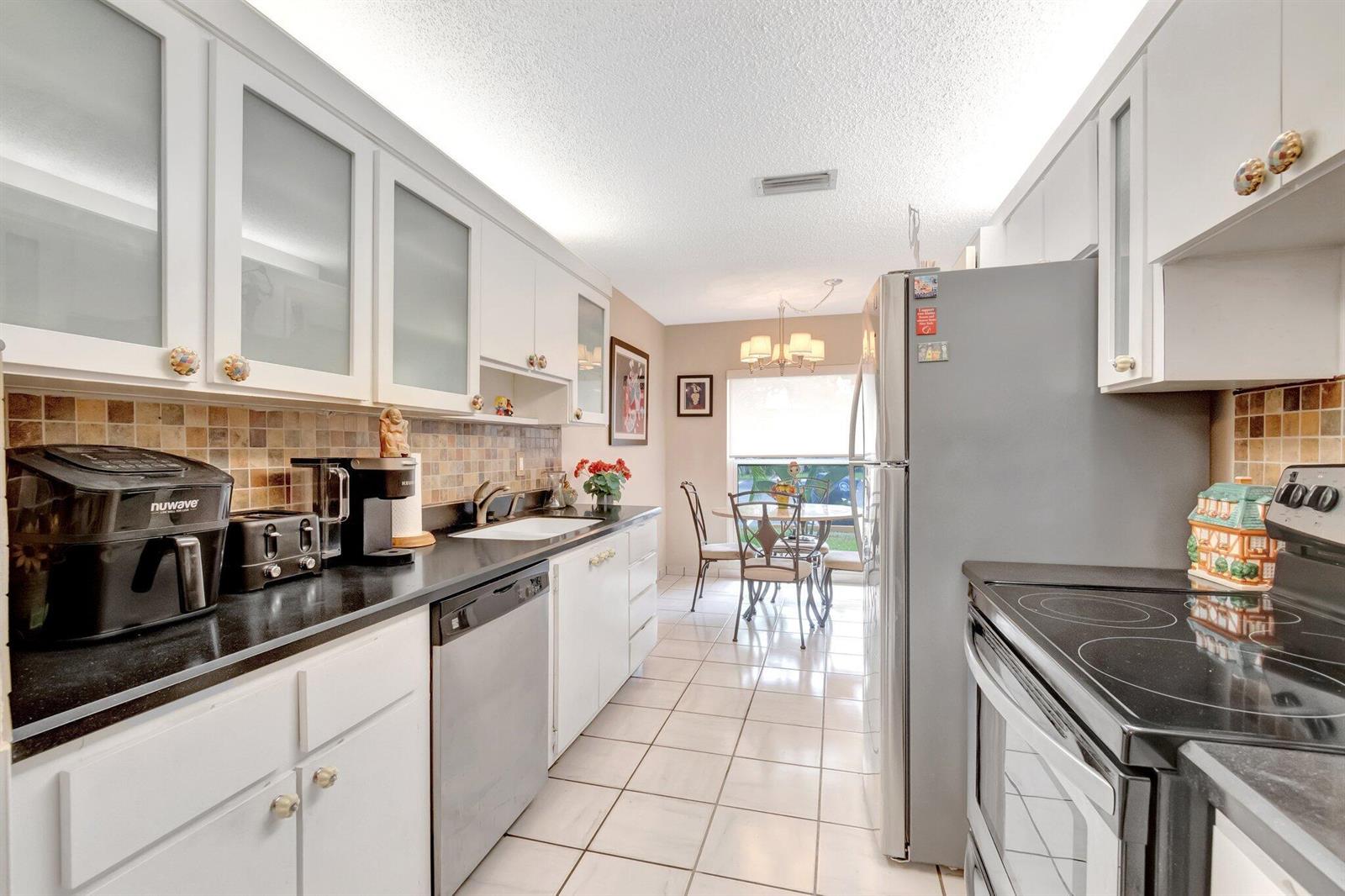 A galley kitchen features white cabinets, black countertops, stainless steel appliances, and tile flooring — 13691 Via Aurora, Delray Beach FL 33484, DELRAY BEACH, FL