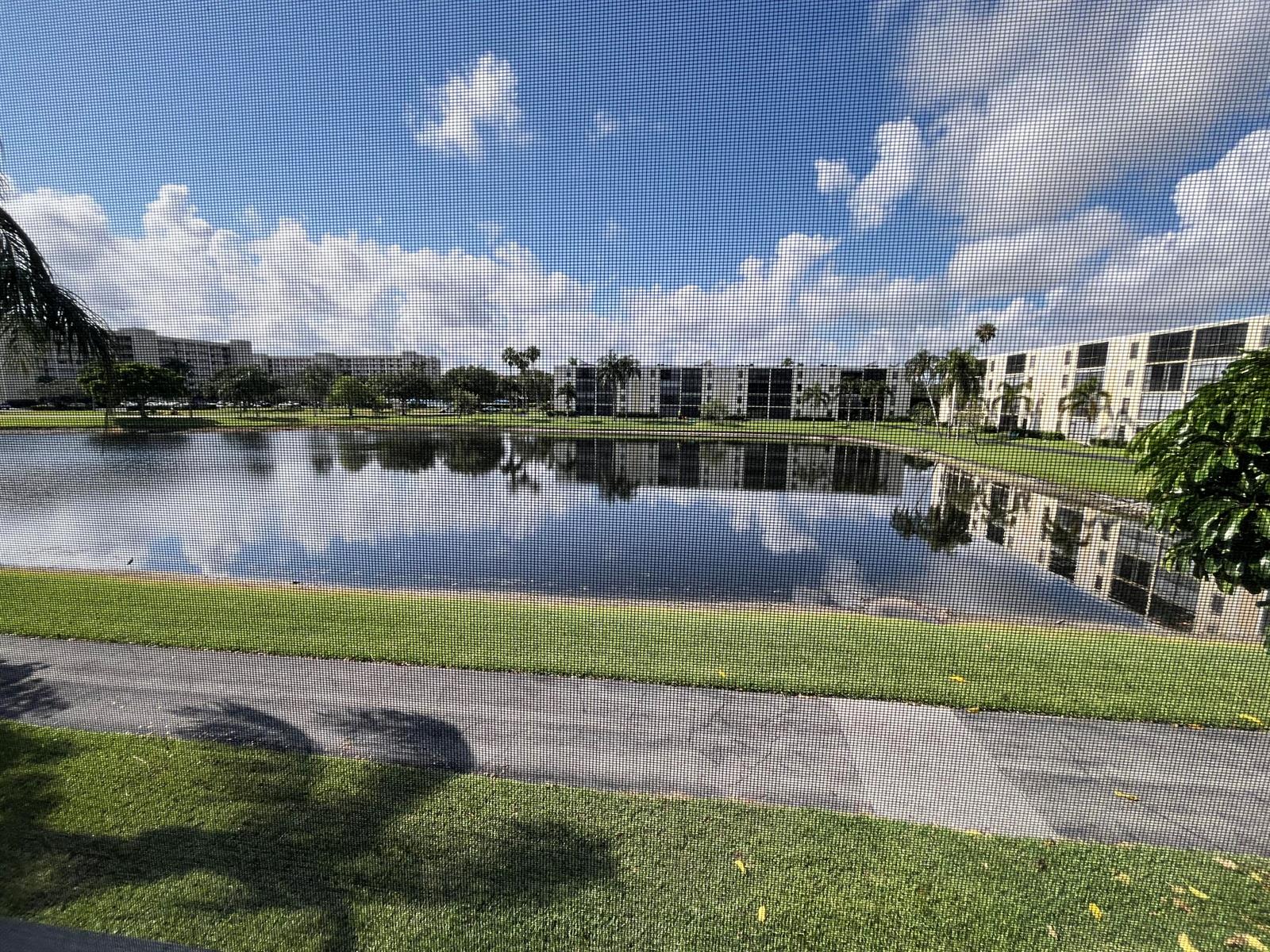 A view of a lake and apartment buildings seen through a screened-in porch window — 7340 Amberly Lane # 205, Delray Beach FL 33446, DELRAY BEACH, FL