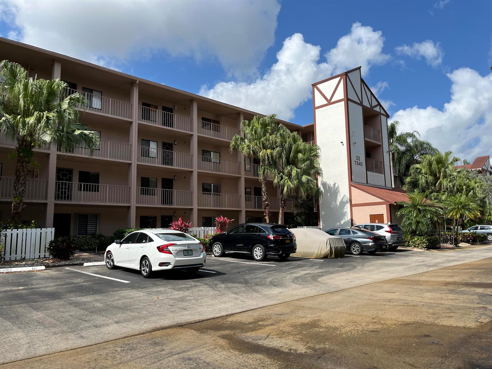 A multi-story apartment building with a parking lot and palm trees under a blue sky — 7340 Amberly Lane # 205, Delray Beach FL 33446, DELRAY BEACH, FL