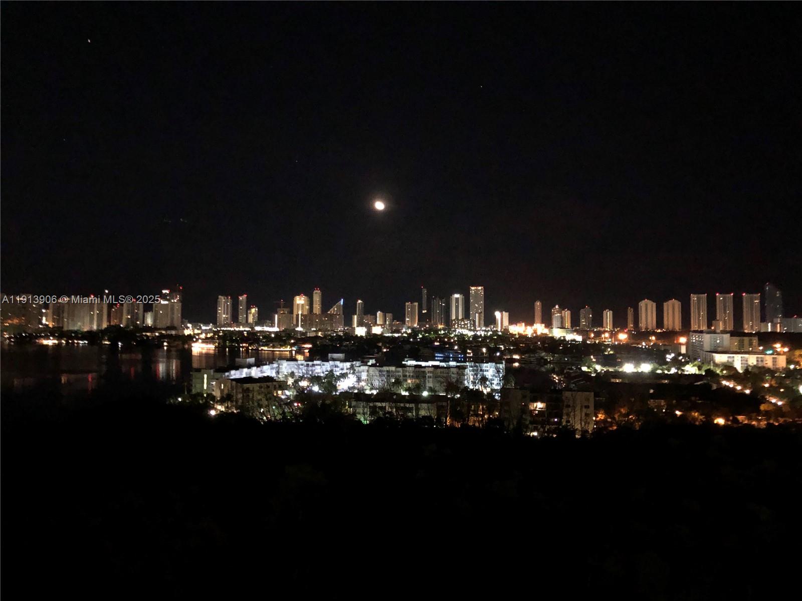 A night view of a city skyline featuring illuminated high-rise buildings and a full moon — 16385 Biscayne Blvd # 3003, North Miami Beach FL 33160, NORTH MIAMI BEACH, FL