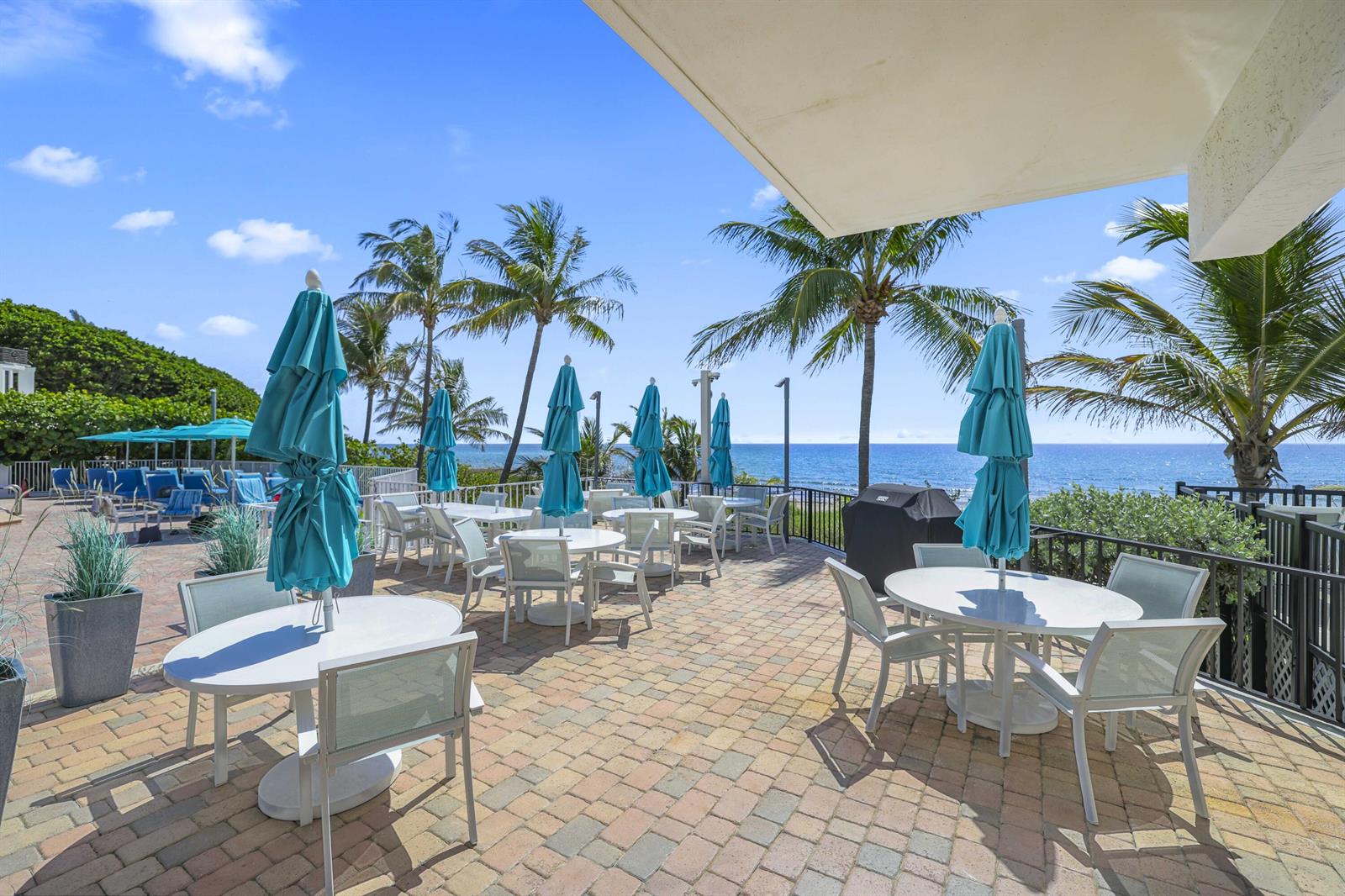 An outdoor patio features multiple dining tables with blue umbrellas overlooking the ocean — 4748 S Ocean Boulevard # 502, Highland Beach FL 33487, HIGHLAND BEACH, FL