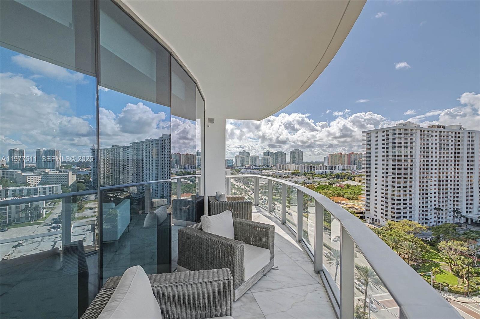 A curved balcony with wicker chairs overlooks a city skyline and a large apartment building — 17475 Collins Ave # 904, Miami Beach FL 33160, MIAMI BEACH, FL