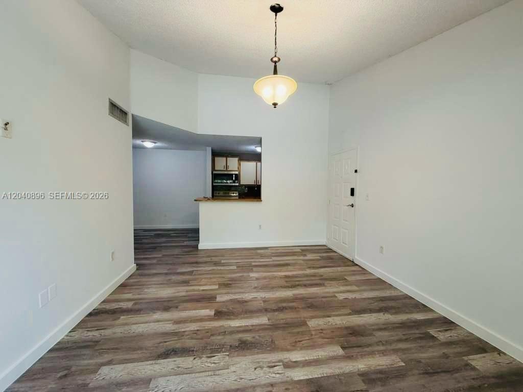 An empty room features wood-look flooring, white walls, a hanging light fixture, and a kitchen — 15530 SW 80th St # 306, Miami FL 33193, MIAMI, FL