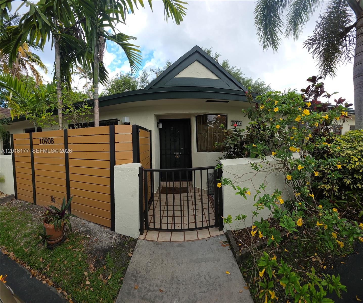A small house exterior features a gated courtyard, a wooden privacy fence, and yellow flowers — 10908 SW 72nd St # 215, Miami FL 33173, MIAMI, FL