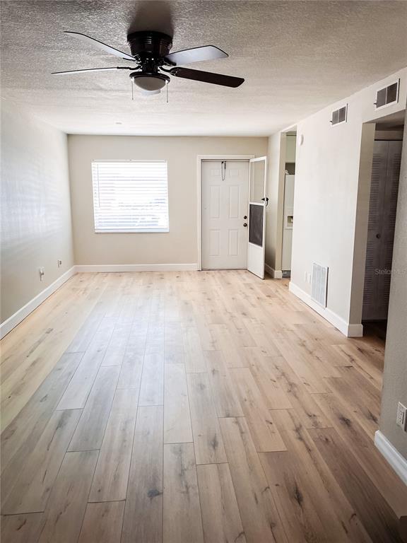 An empty living room features light wood-look flooring, a ceiling fan, and a white door — 24862 US HIGHWAY 19 N UNIT 1602, CLEARWATER, FL