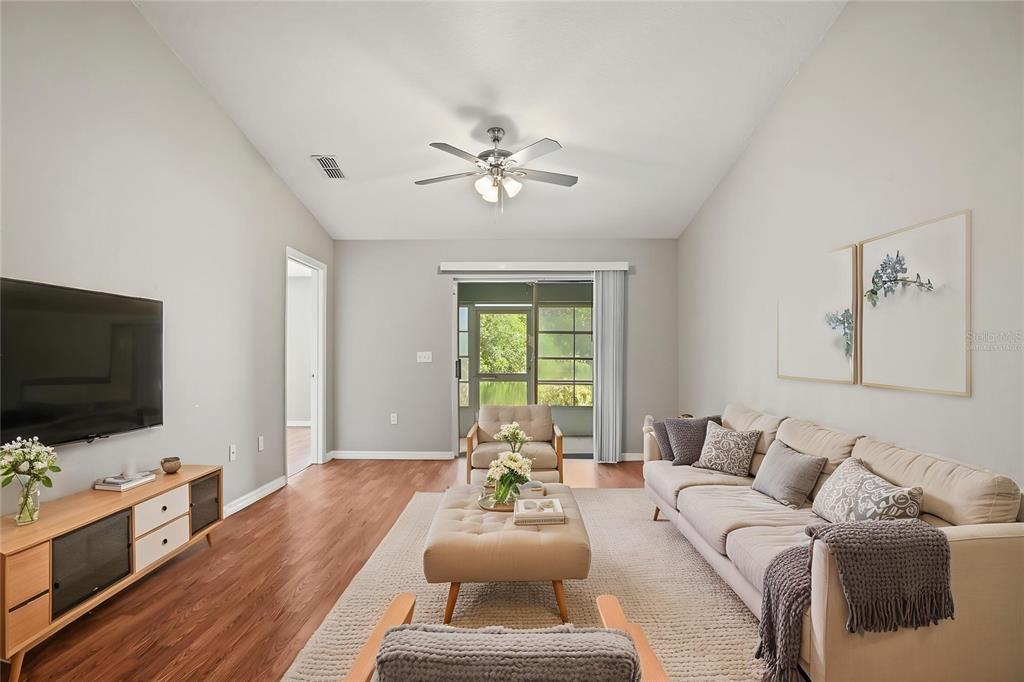A living room features a beige sofa, a tufted ottoman, wood flooring, and a ceiling fan — 3435 VILLAGE GREEN COURT, ST CLOUD, FL