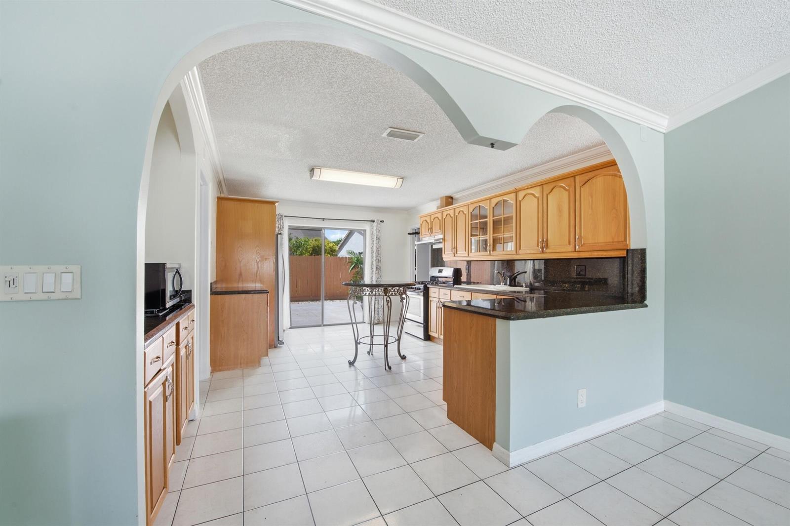 A kitchen with light wood cabinets, dark countertops, and white tile flooring under an archway — 5814 Ithaca Circle E, Lake Worth FL 33463, LAKE WORTH, FL