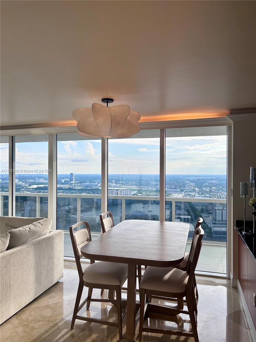 A dining area with a wooden table and chairs sits before floor-to-ceiling windows overlooking a city — 1830 S Ocean Dr # 4106, Hallandale Beach FL 33009, HALLANDALE BEACH, FL