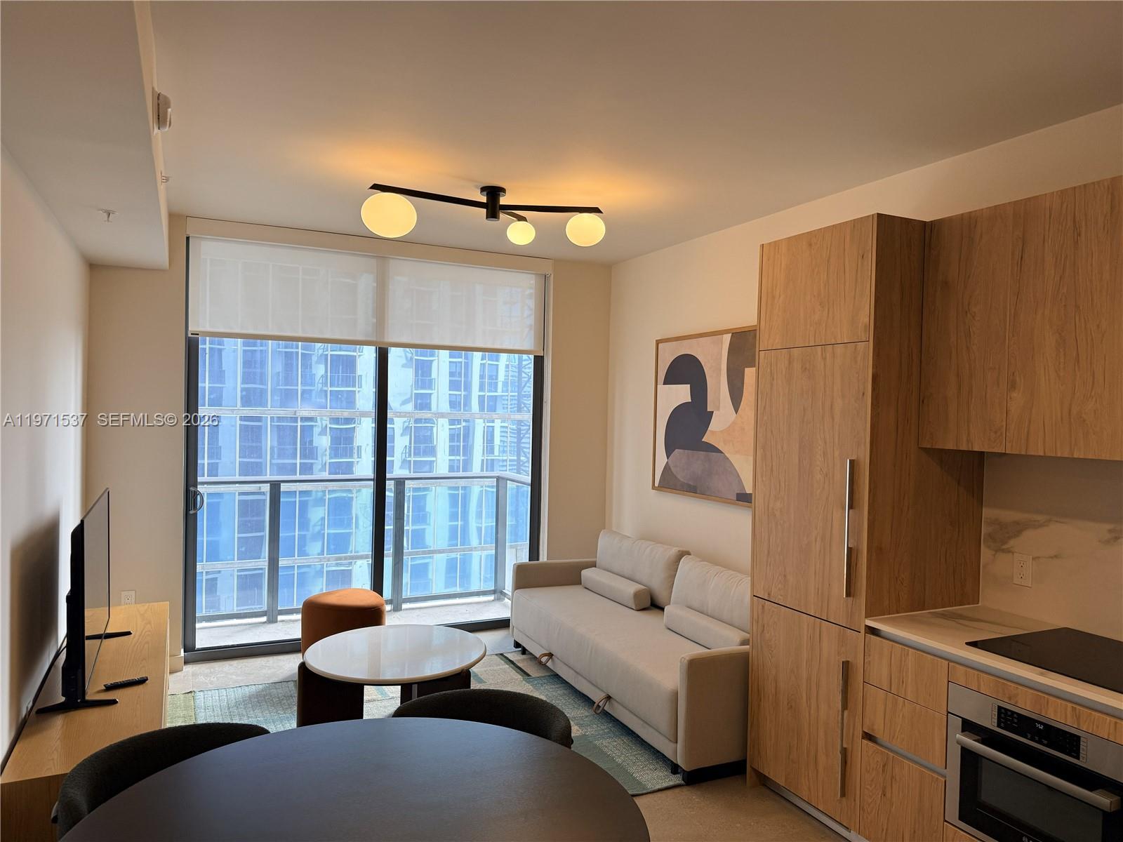 A modern living room and kitchen area features light wood cabinetry and a balcony view — 11 NE 6th # 1905, Miami FL 33132, MIAMI, FL