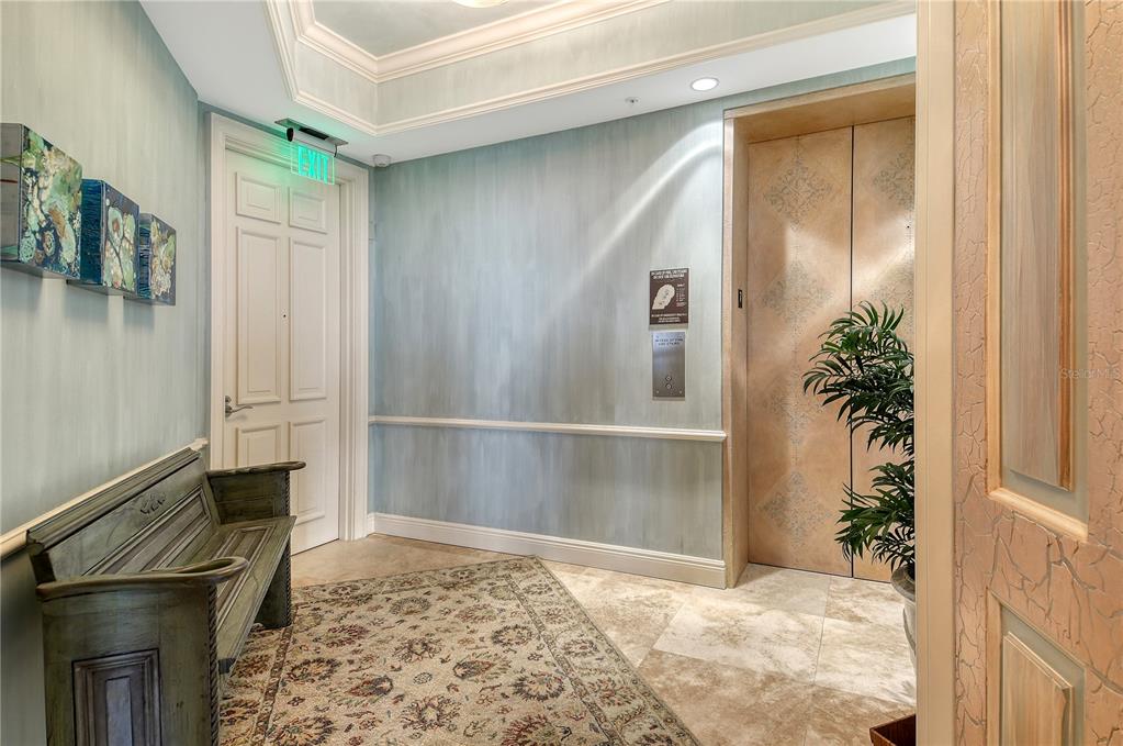 An elevator lobby features light blue walls, a patterned rug, a wooden bench, and tile flooring — 1300 BENJAMIN FRANKLIN DRIVE UNIT 709, SARASOTA, FL