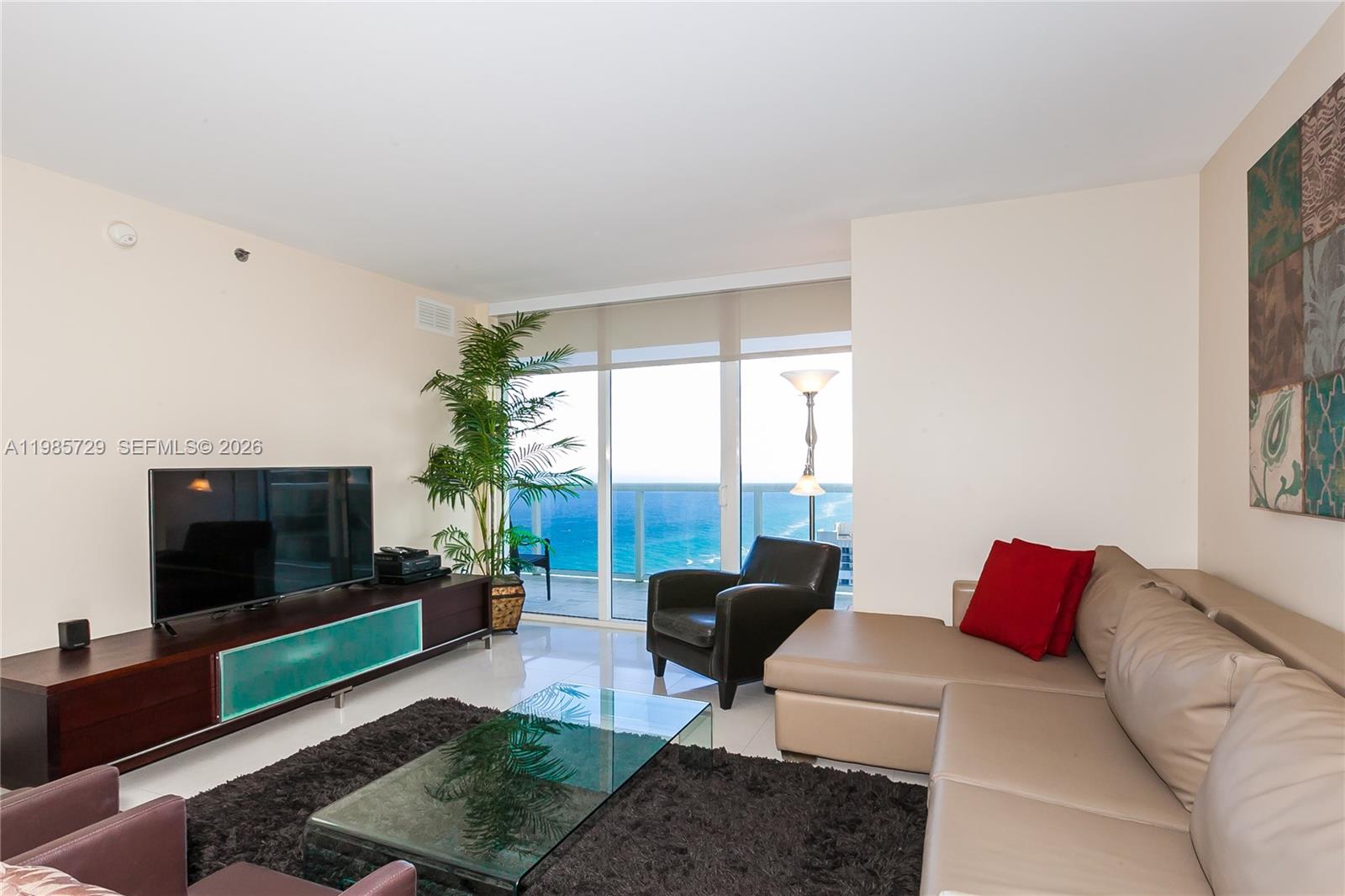 A living room features a sectional sofa, glass coffee table, television, and ocean view balcony — 1830 S OCEAN DR # 3204, Hallandale Beach FL 33009, HALLANDALE BEACH, FL