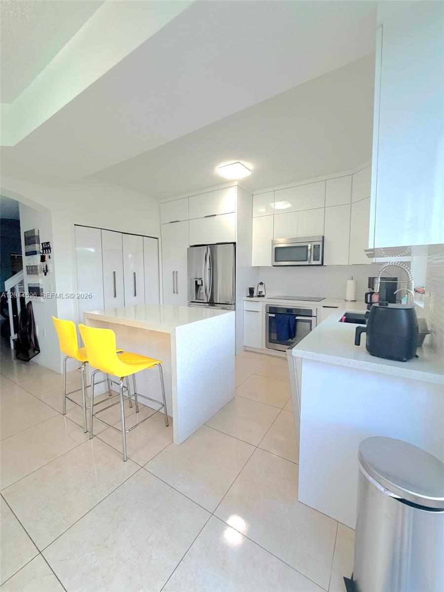 A modern kitchen features white cabinetry, a central island with yellow stools, and tile flooring — 11405 NW 62nd Ter # 236, Doral FL 33178, DORAL, FL