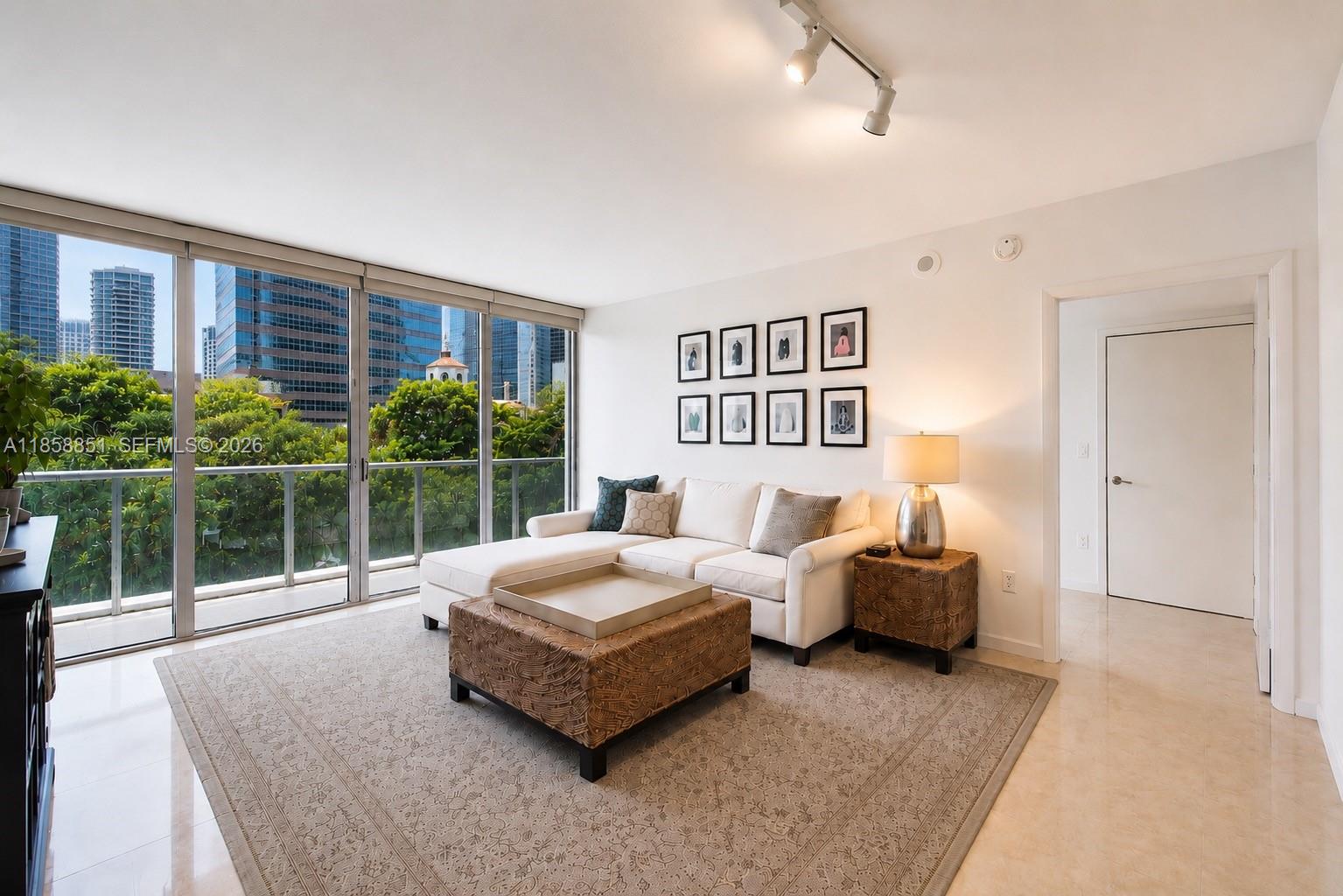 A living room features a white sectional sofa, patterned ottoman, and floor-to-ceiling windows overlooking buildings — 495 Brickell Ave # 411, Miami FL 33131, MIAMI, FL