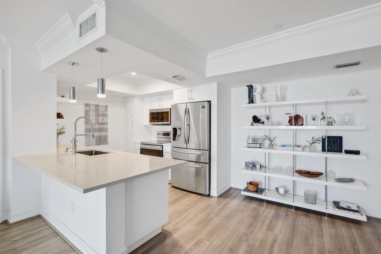 A modern kitchen features a white island, stainless steel appliances, and open shelving on white walls — 99 SE Mizner Boulevard # 609, Boca Raton FL 33432, BOCA RATON, FL