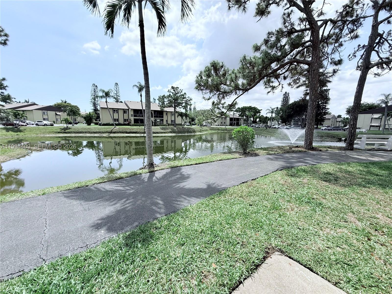 A paved path winds alongside a pond with a fountain near apartment buildings and trees — 340 Pine Ridge Cir # 1, Green Acres FL 33463, GREEN ACRES, FL