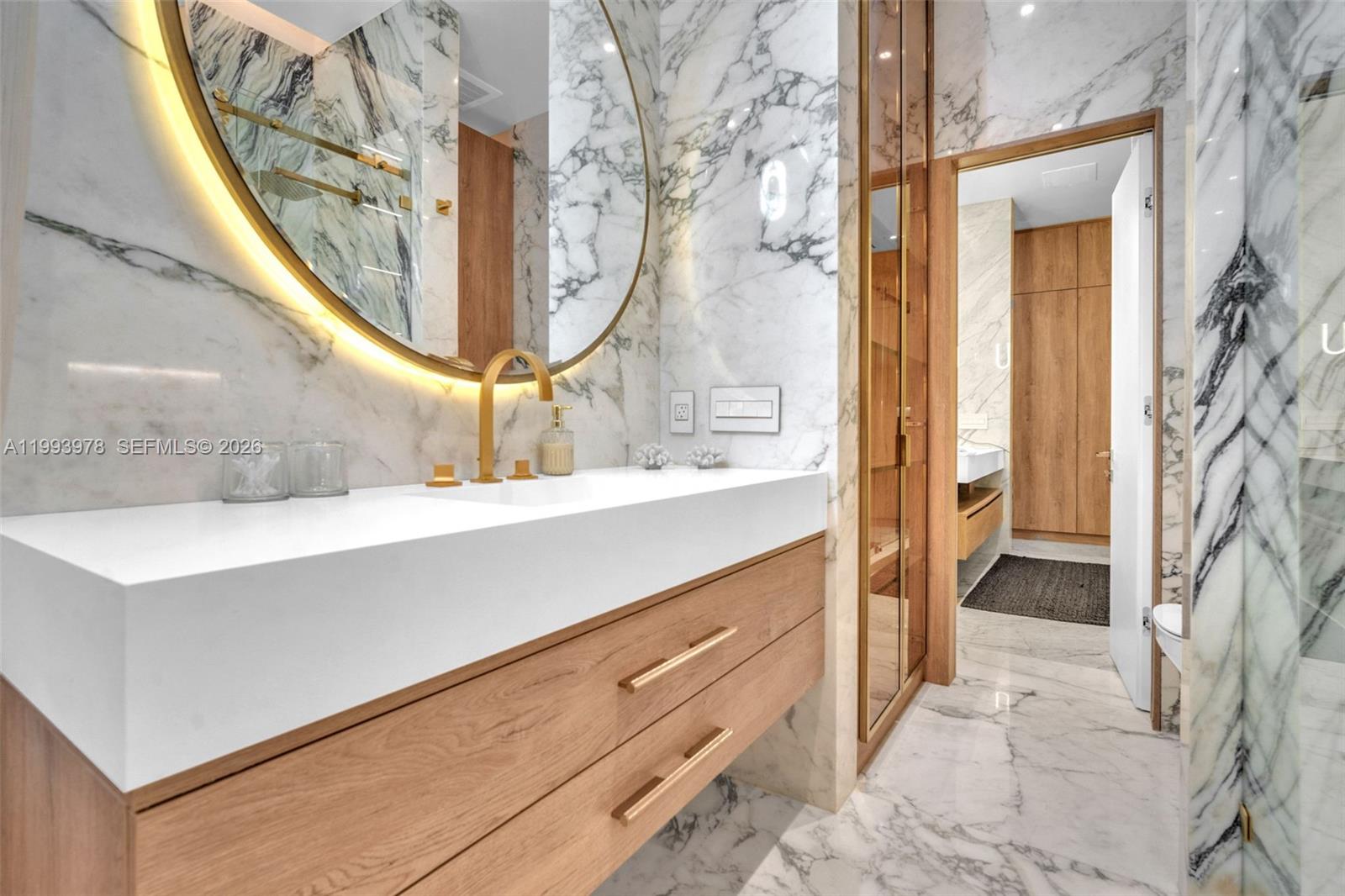 A modern bathroom features a floating wood vanity, marble walls, and a circular backlit mirror — 46 NE 162nd St, Miami FL 33162, MIAMI, FL