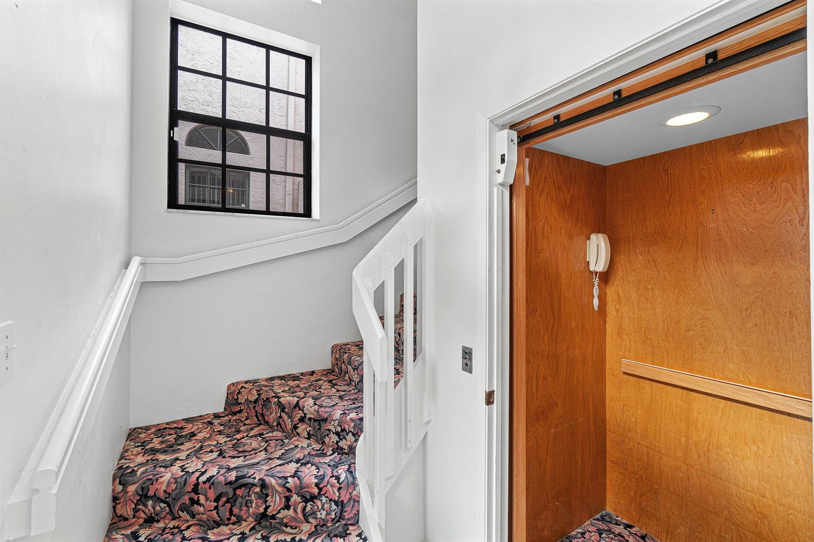 A staircase with floral carpet sits beside a small residential elevator with wood-paneled walls — 5171 Europa Drive # G, Boynton Beach FL 33437, BOYNTON BEACH, FL