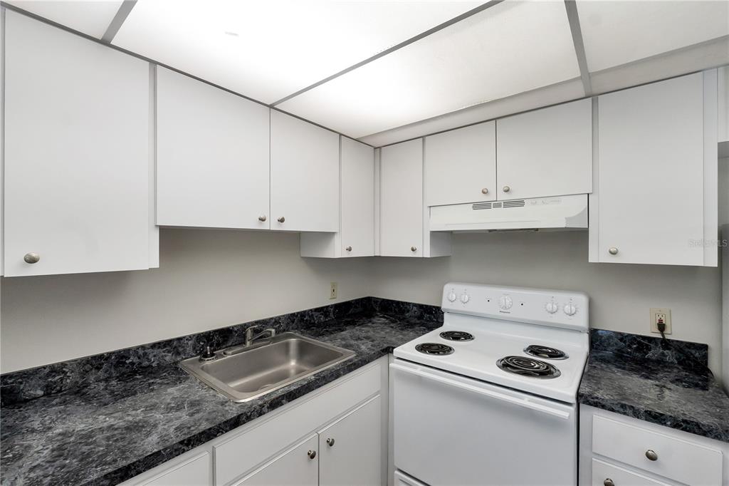 A small kitchen features white cabinets, dark marble-patterned countertops, and a white electric stove — 201 KINGS BOULEVARD UNIT 5, SUN CITY CENTER, FL