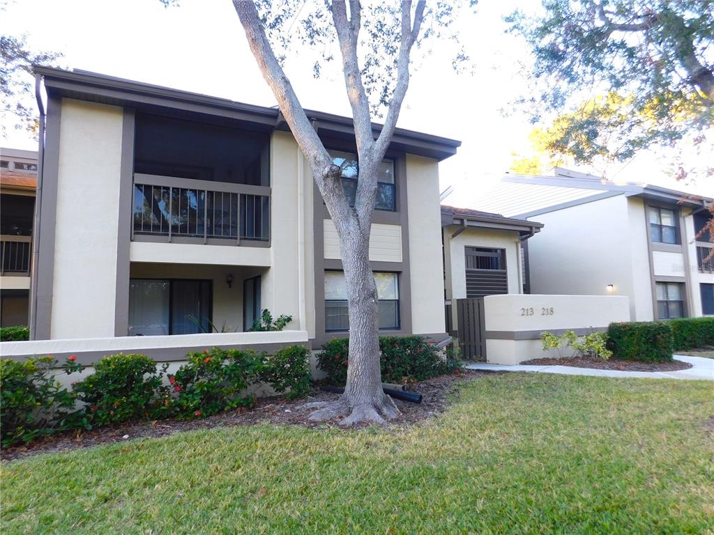 A two-story apartment building exterior features tan stucco walls, brown trim, and a screened balcony — 218 WOODLAKE WYNDE UNIT 218, OLDSMAR, FL