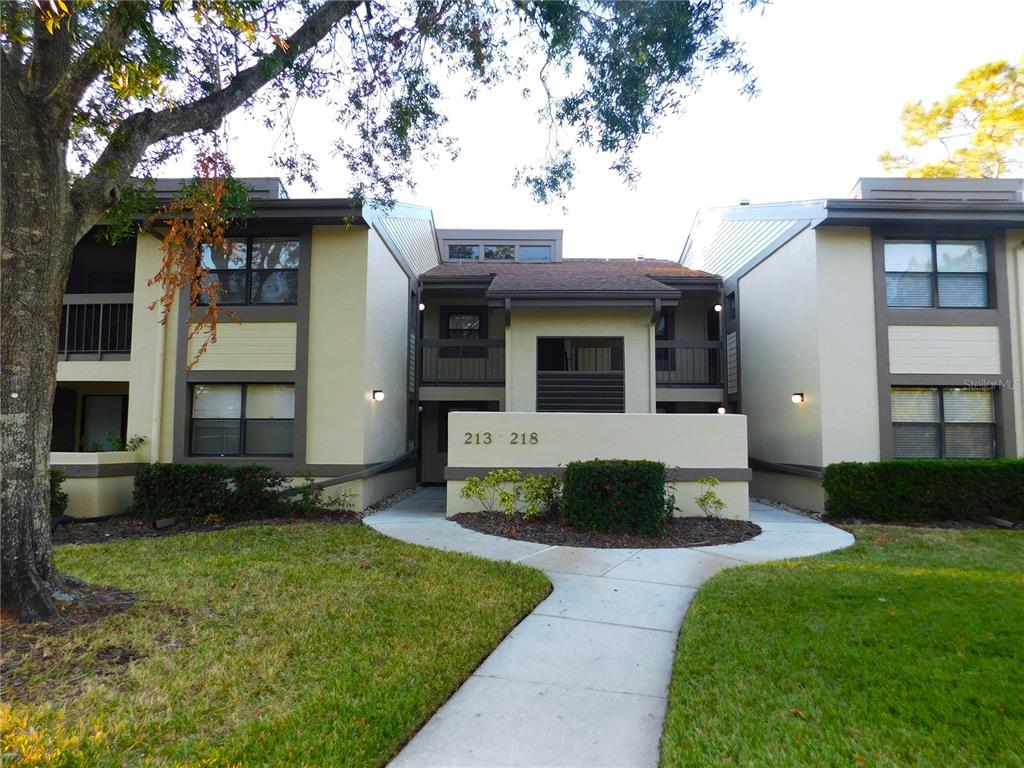 A two-story apartment building exterior features tan stucco walls, brown trim, and a concrete walkway — 218 WOODLAKE WYNDE UNIT 218, OLDSMAR, FL