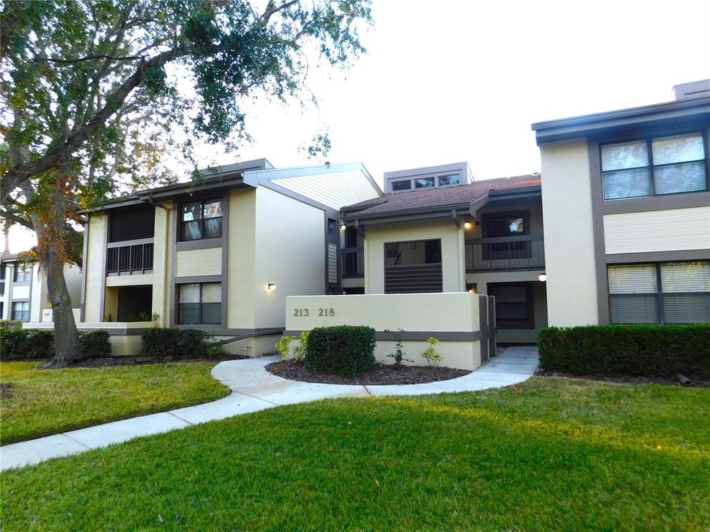 A two-story apartment building exterior features tan siding, dark trim, and a concrete walkway — 218 WOODLAKE WYNDE UNIT 218, OLDSMAR, FL