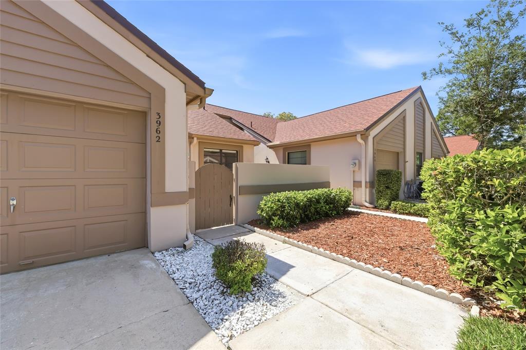 A tan house exterior features a garage, a gated courtyard entrance, and a landscaped yard — 3962 MERMOOR DRIVE, PALM HARBOR, FL