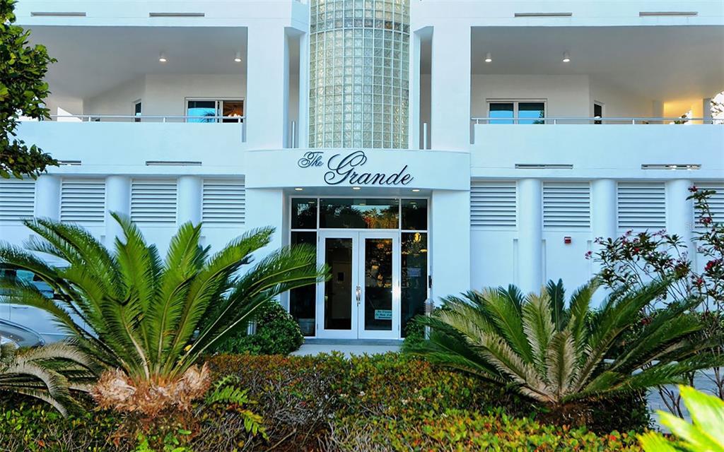 The exterior entrance of a white building named The Grande features glass doors and landscaping — 4561 GULF OF MEXICO DRIVE UNIT 202, LONGBOAT KEY, FL