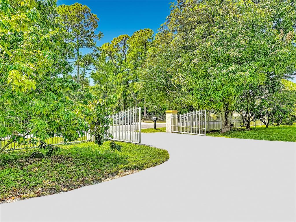 A white metal gate opens onto a paved driveway surrounded by lush green trees — 12821 86TH ROAD N, WEST PALM BEACH, FL