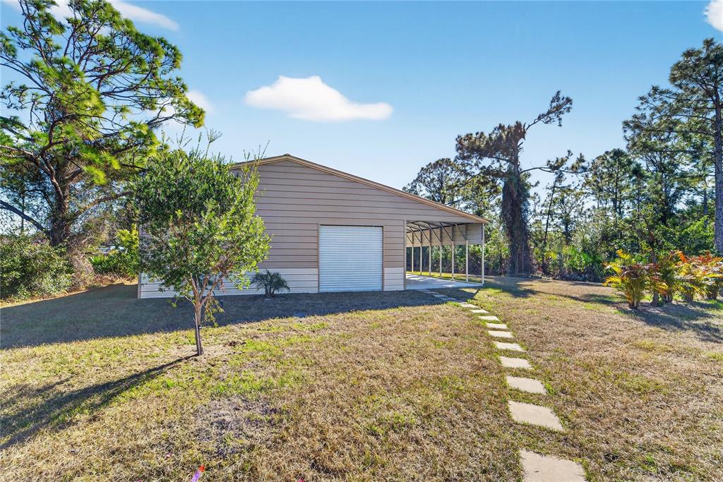 A tan metal garage with a side carport stands on a grassy lot with trees — 7888 EVINRUDE AVENUE, NORTH PORT, FL