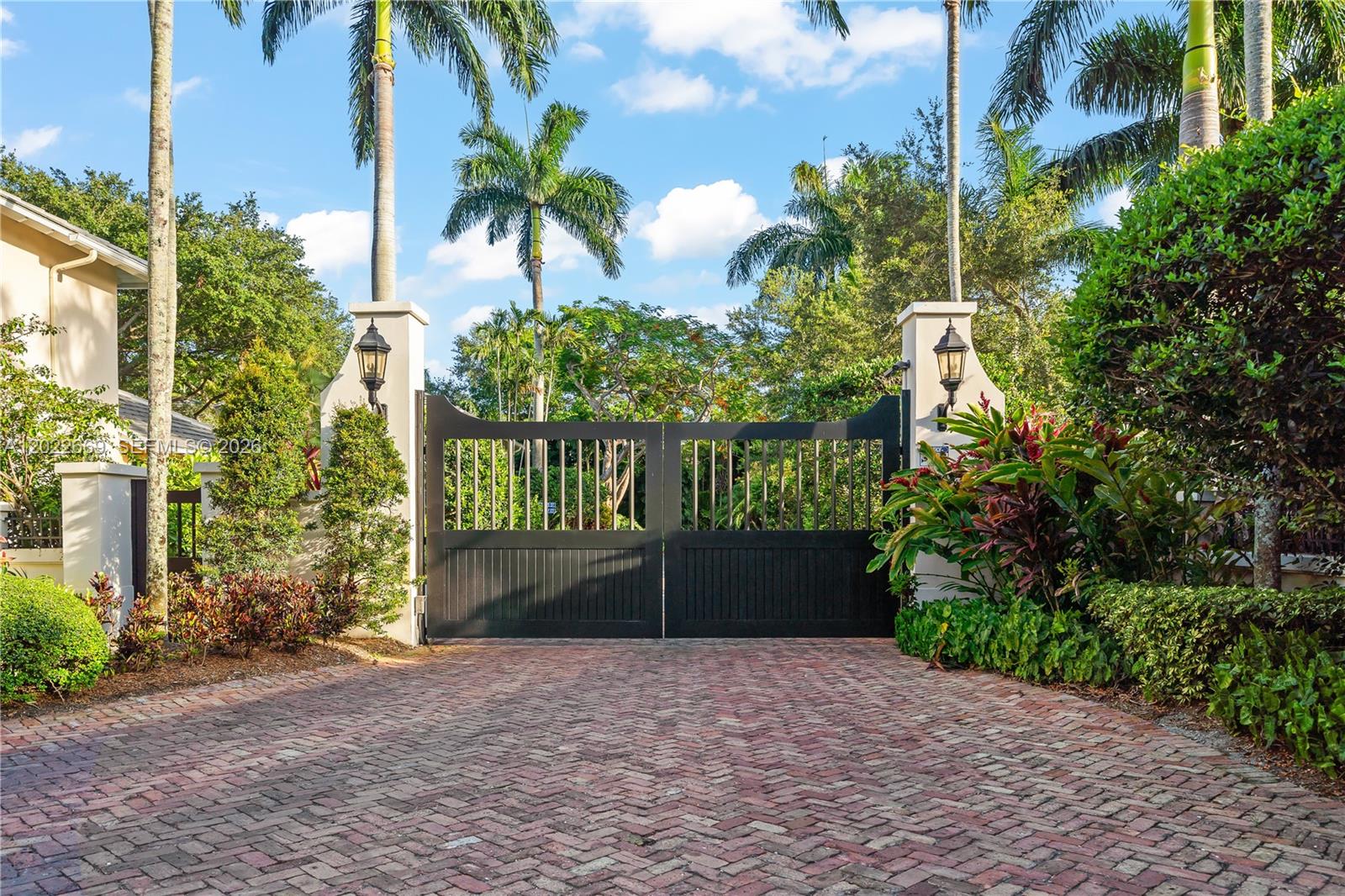A black metal gate stands between two stucco pillars at the end of a brick driveway — 5700 SW 86th St, South Miami FL 33143, SOUTH MIAMI, FL