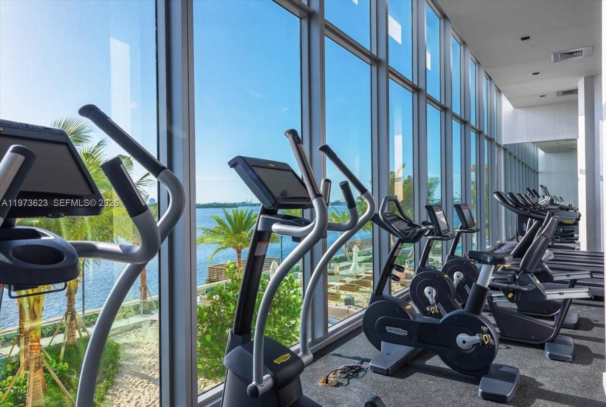 A gym features rows of exercise equipment facing floor-to-ceiling windows overlooking a waterfront area — 2900 NE 7th Ave # 3004, Miami FL 33137, MIAMI, FL